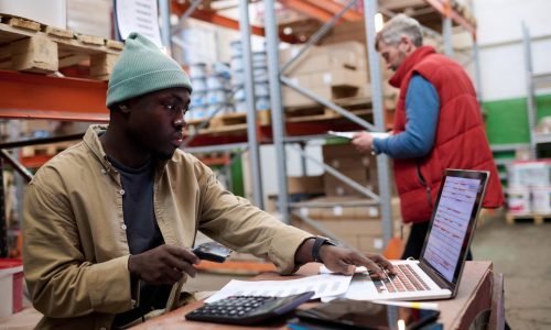 African American distributor using laptop to scan barcodes of parcels while sitting at his workplace at warehouse